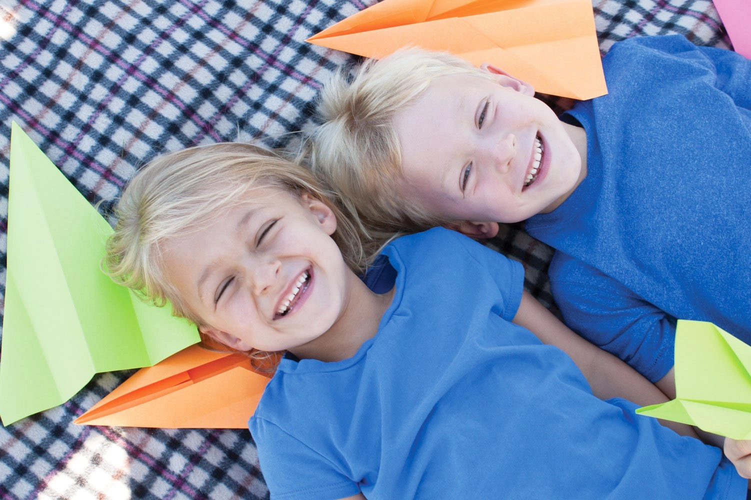 A smiling young boy and girl laying on a blanket, playing with paper planes.